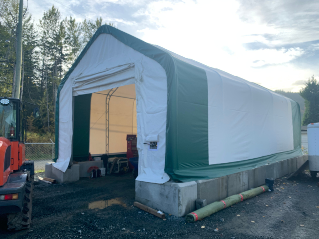 A white and green PVC fabric garage structure with an open door, built on a concrete foundation. The interior is visible, showing storage equipment.