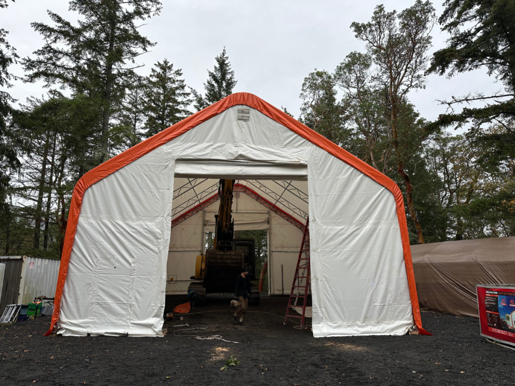 A heavy-duty fabric garage shelter with an open roll-up door, revealing a large excavator inside, demonstrating the shelter’s spacious and high-clearance design for heavy equipment storage.